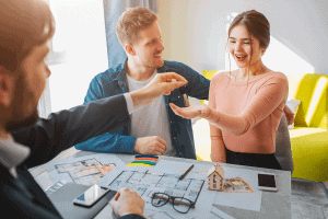 A couple sits across a table from a Portland real estate agent handing them a key. Blueprints and a small model house are visible on the table. The woman smiles excitedly while the man looks on happily. Bright sunlight fills the room.