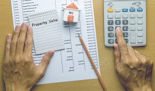 Hands are seen using a calculator on a wooden desk with financial documents. A notepad labeled Property Value and a paper house resting on a pencil are also visible, perfect for anyone seeking Portland real estate advice.