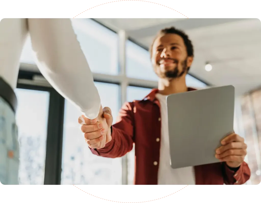Two people are shaking hands indoors. One person, holding a tablet, wears a red shirt. They are smiling at each other, suggesting a friendly or business interaction. Bright natural light filters through large windows in the background.
