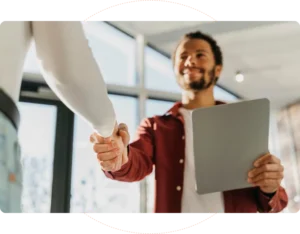 Two people are shaking hands indoors. One person, holding a tablet, wears a red shirt. They are smiling at each other, suggesting a friendly or business interaction. Bright natural light filters through large windows in the background.
