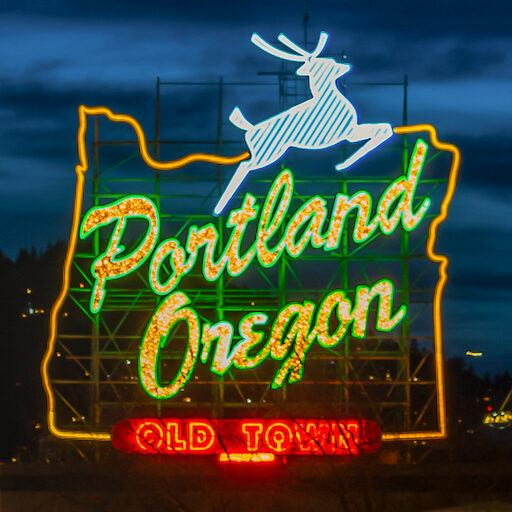 Neon sign reading Portland Oregon with a white outline of the state shape and a leaping stag at the top. The words Old Town are lit in red at the bottom of the sign. The background is a dusk sky.